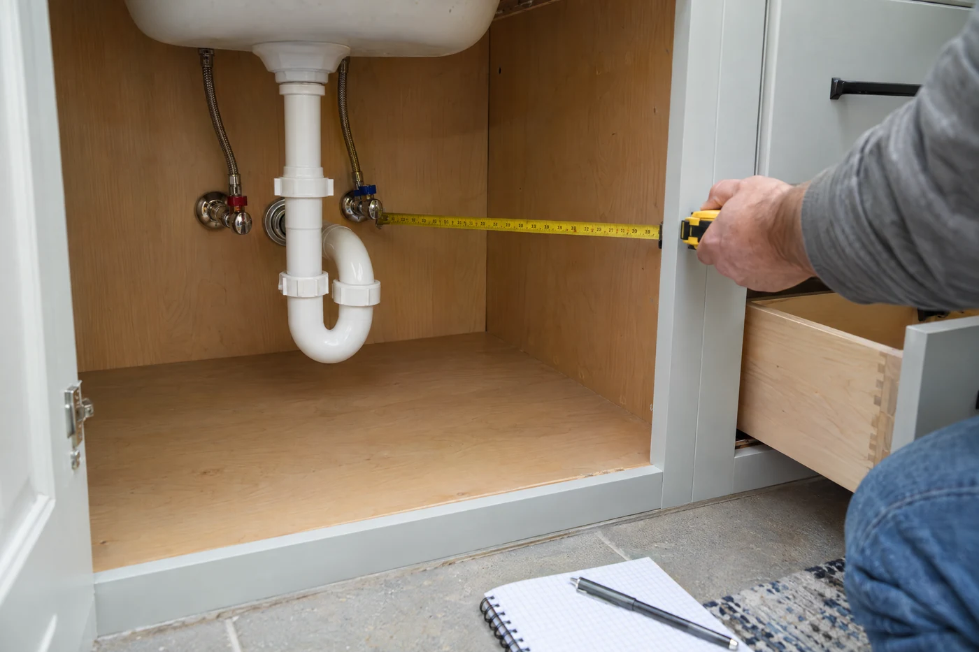 Illustrative view inside a vanity cabinet showing a drain trap, shutoff valves, a tape measure, and an open drawer